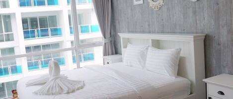 Une chambre moderne avec un grand lit, une table de chevet, une commode et une vue sur un immeuble par la fenêtre.