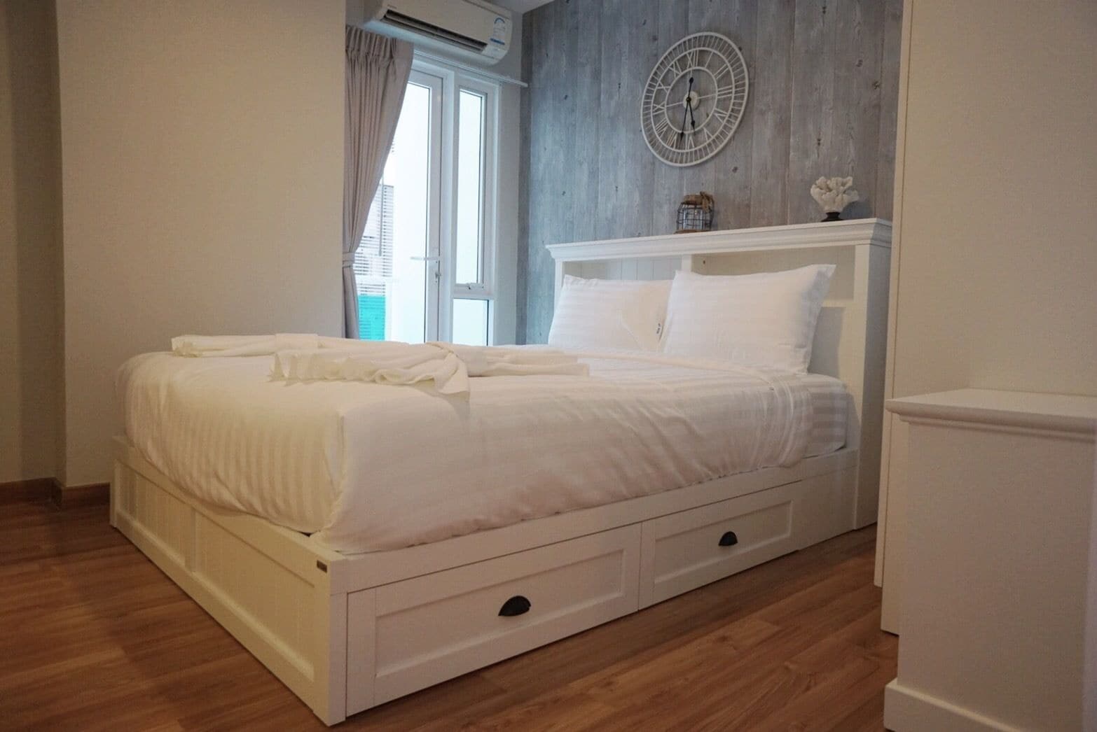 Une chambre à coucher avec un lit blanc, un plancher en bois, une fenêtre avec des rideaux et une horloge murale.