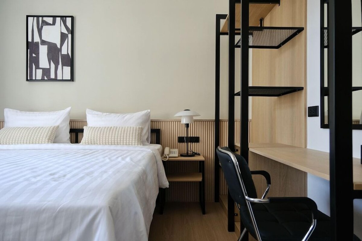 A hotel room with a bed, a desk, a chair, a lamp, and a wall-mounted shelf.