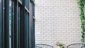 Un coin de pièce avec un mur en briques, deux chaises noires en fil de fer, une petite table avec une plante et un seau en métal contenant des ustensiles.