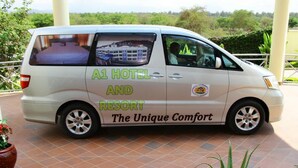 Una camioneta blanca con la marca "A1 HOTEL AND RESORT The Unique Comfort", estacionada sobre una superficie de baldosas y con follaje verde al fondo.