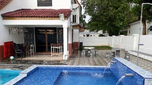 A swimming pool with a tiled deck, a covered patio area with chairs and tables, and a white fence in the background.