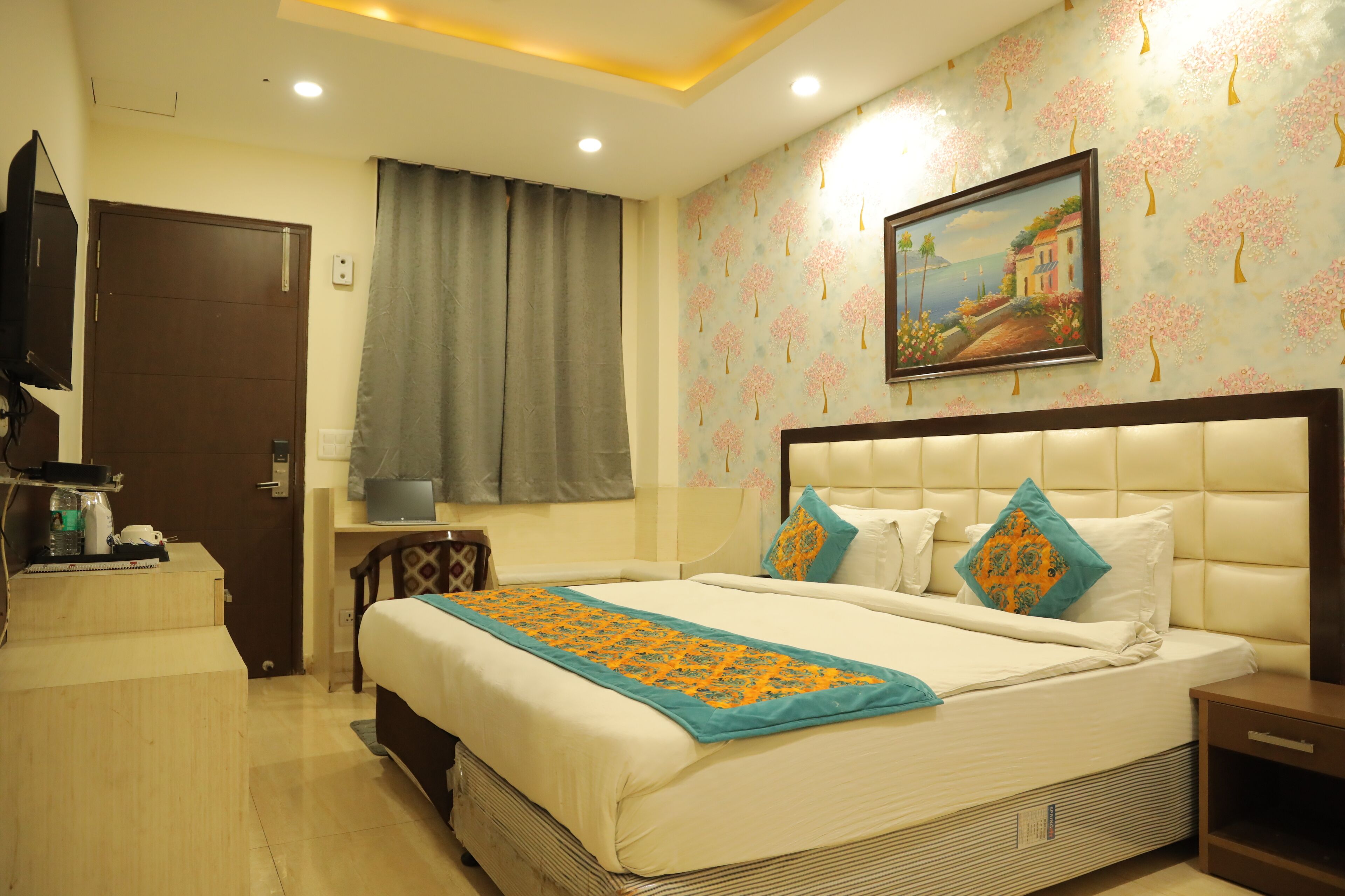 A hotel room with a bed, a nightstand, a desk with a lamp, a painting on the wall, and a window with curtains.