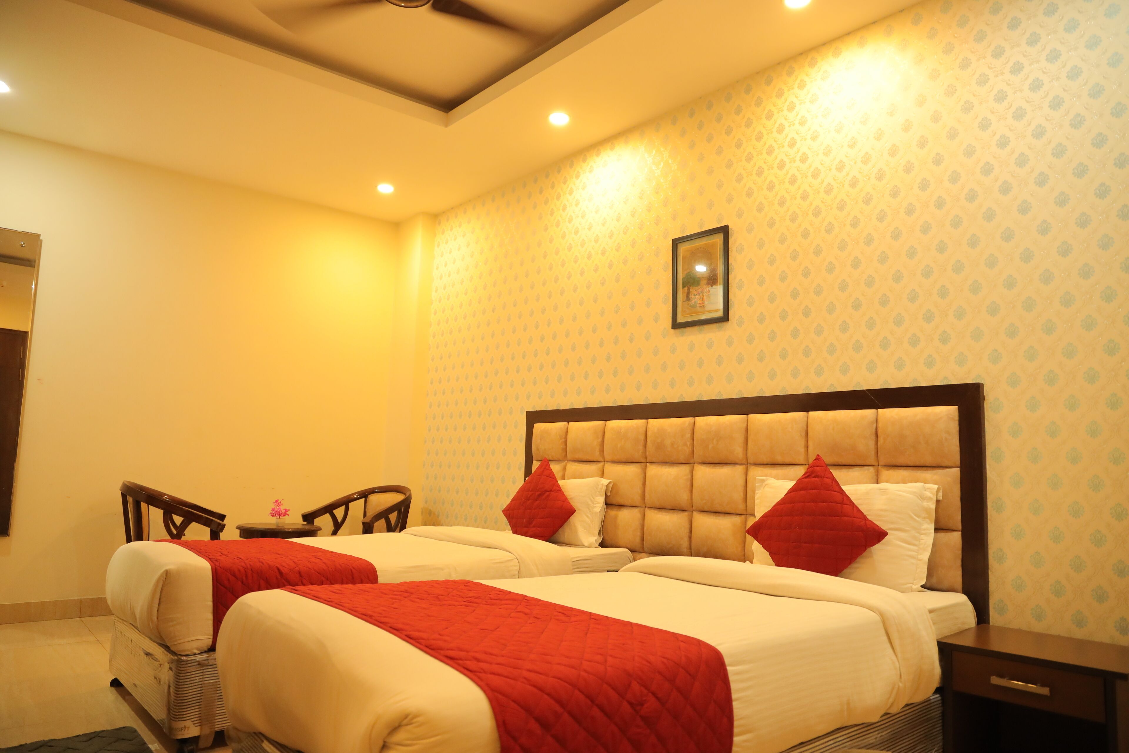 A hotel room with two beds, each with a red blanket and pillows, a wooden headboard, a small table with a chair, and a framed picture on the wall.