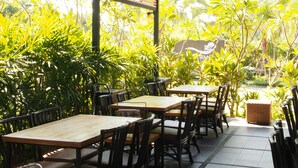 An outdoor seating area with wooden tables and chairs, surrounded by lush greenery and a view of a building with a logo. - Panwana Resort (Rayong)