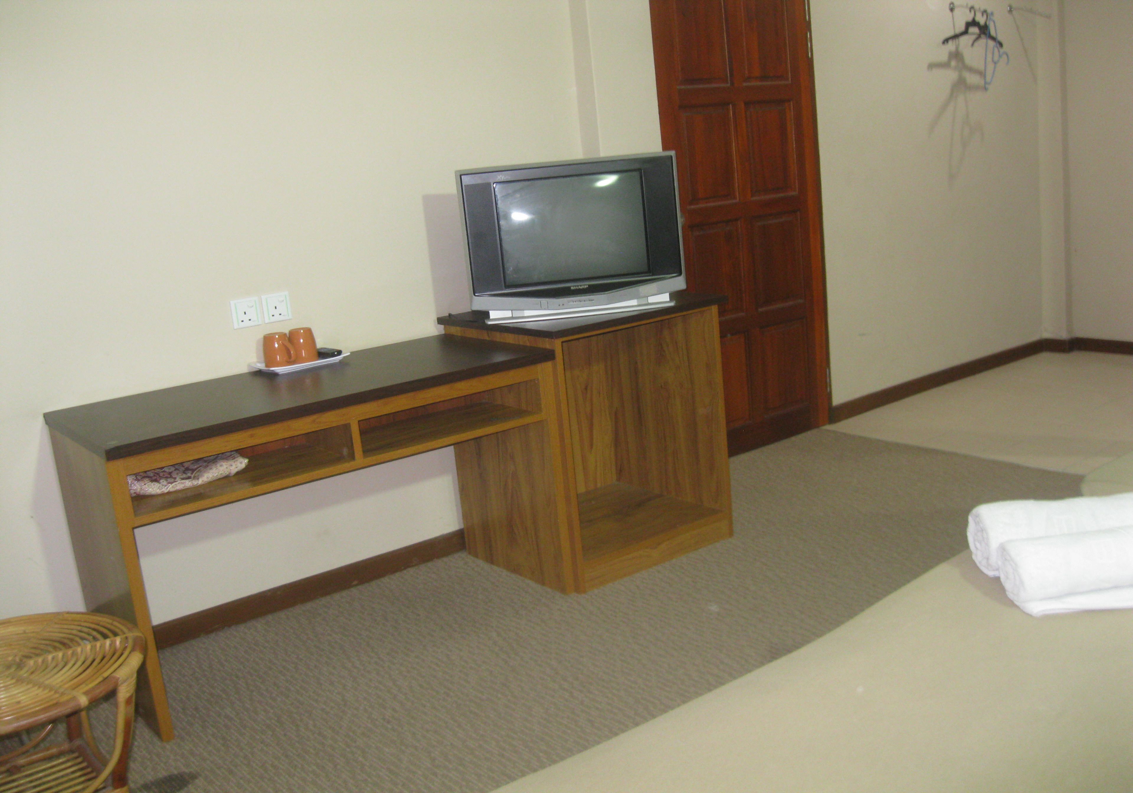 Ein kleines Zimmer mit Schreibtisch aus Holz, einem Fernseher darauf, einem Sessel und einem Bett mit weißem Handtuch.