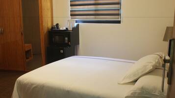 A hotel room with a bed, a bedside table with a kettle, a microwave, and a window with blinds.