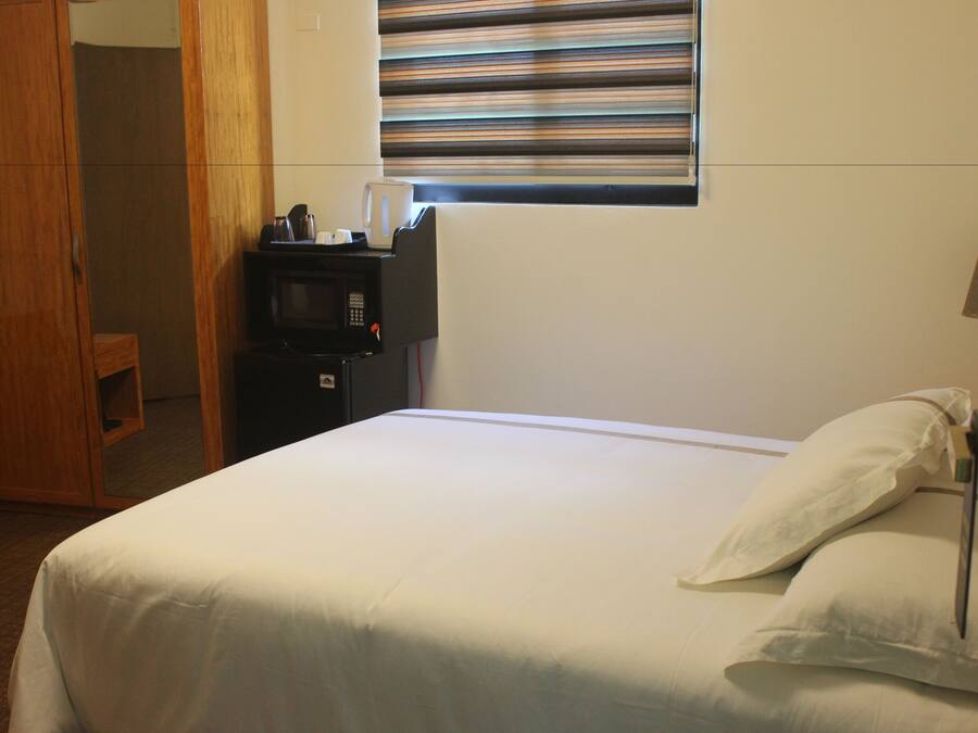 A hotel room with a bed, a bedside table with a kettle, a microwave, and a window with blinds.
