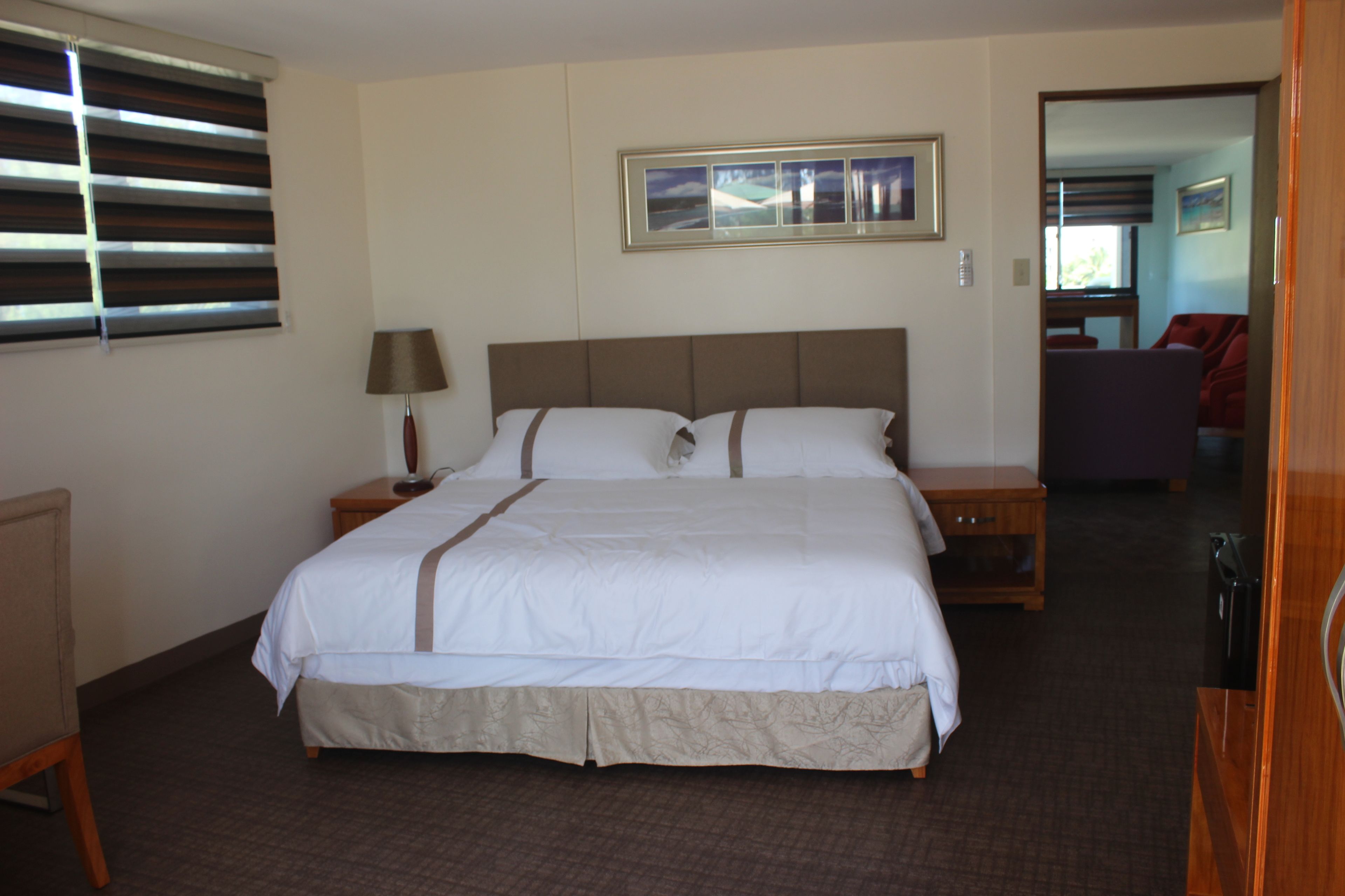 A hotel room with a large bed, a bedside table with a lamp, a framed picture above the bed, and a view of a living area through an open door.