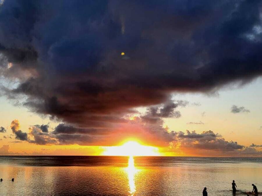 A sunset over a calm sea with a few people in the water.