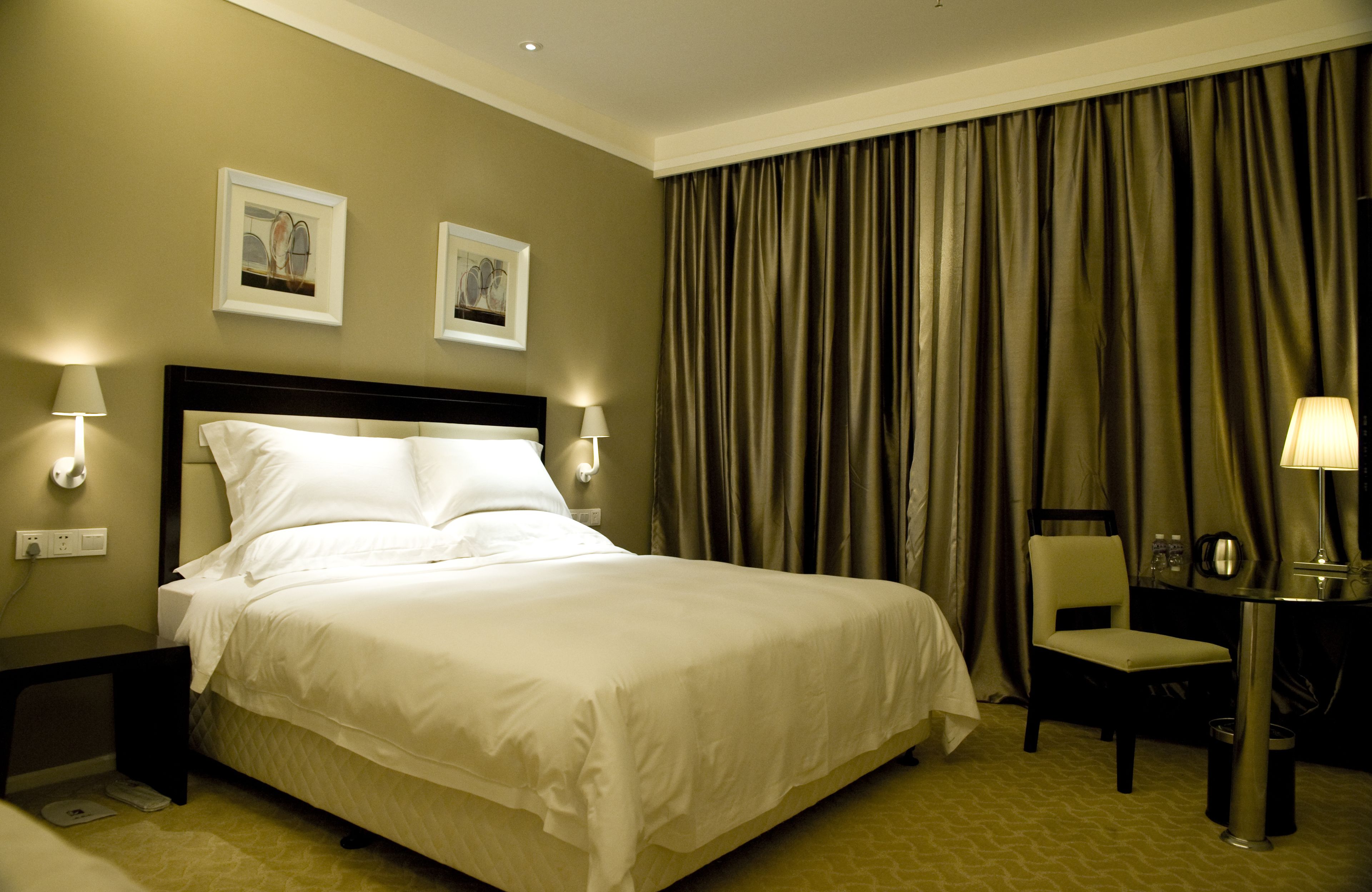 A hotel room with a large bed, two wall-mounted lamps, a desk with a chair, and a large window with curtains.