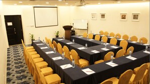 Un salón de conferencias con filas de mesas y sillas, una pantalla de proyector, una pizarra y cuadros en la pared.