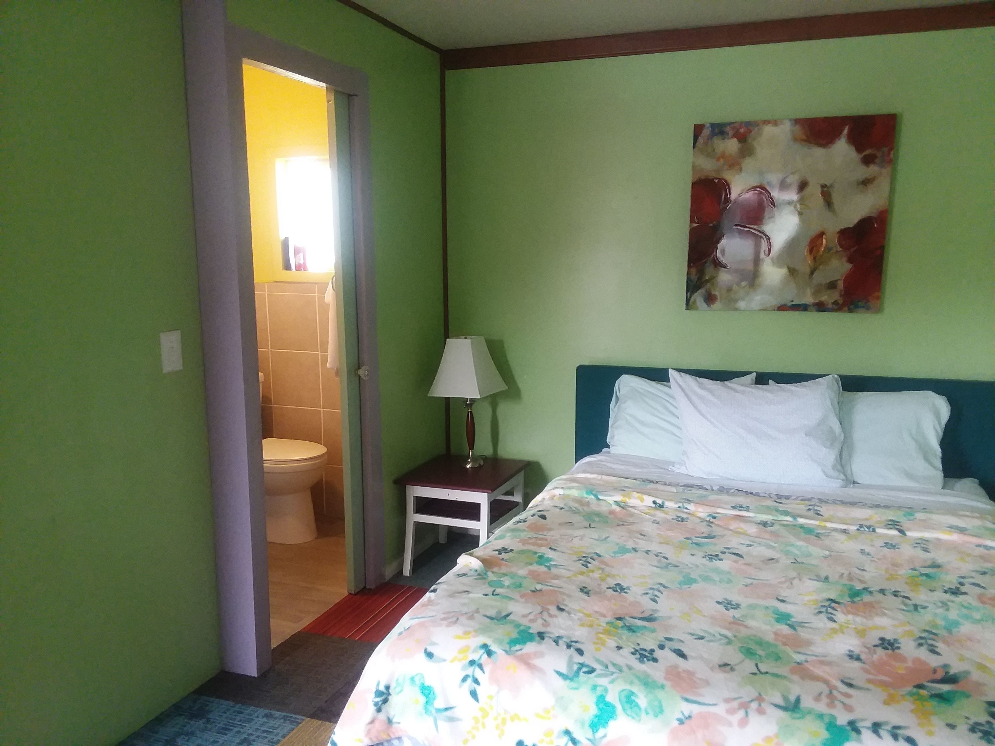 Une chambre d’hôtel avec un lit, une table de chevet, une lampe, un tableau au mur et une salle de bain visible à travers une porte ouverte.