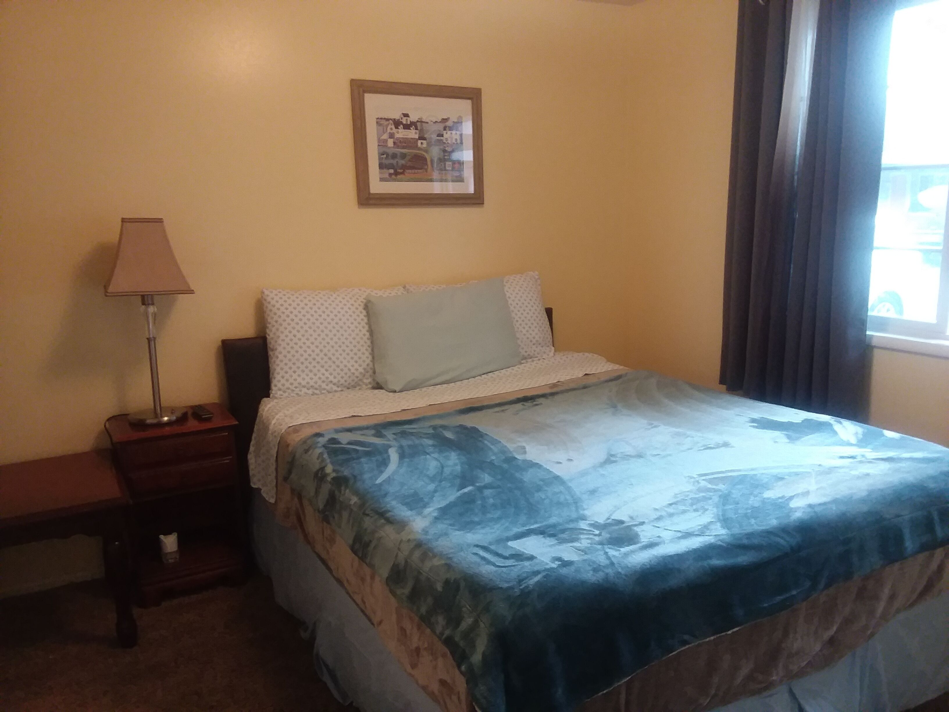 Un dormitorio con cama, mesita de noche, lámpara, un cuadro en la pared y ventana con cortinas.