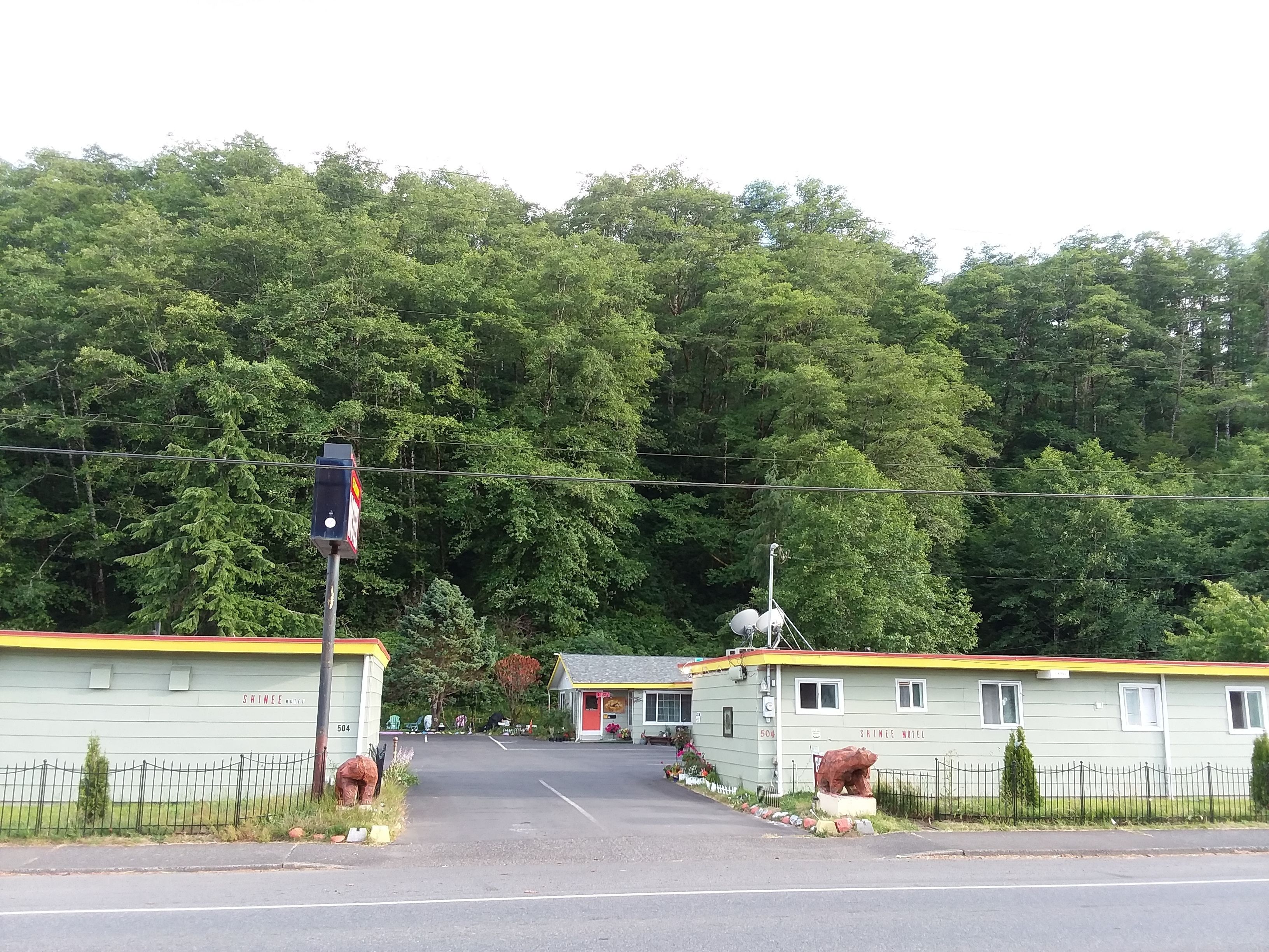 A motel with a simple, functional design, featuring a central road leading to the entrance, surrounded by lush greenery and a dense forest backdrop.
