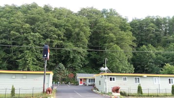 A motel with a simple, functional design, featuring a central road leading to the entrance, surrounded by lush greenery and a dense forest backdrop.
