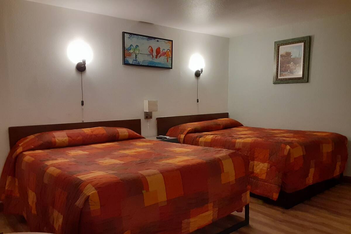 Quarto de hotel com duas camas, cada uma com colcha estampada, uma mesinha de cabeceira, uma luminária na parede e duas fotografias emolduradas.