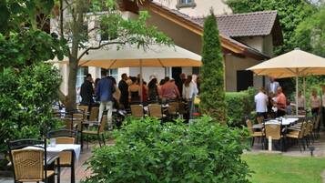 An outdoor dining area with tables and chairs, surrounded by greenery and buildings.