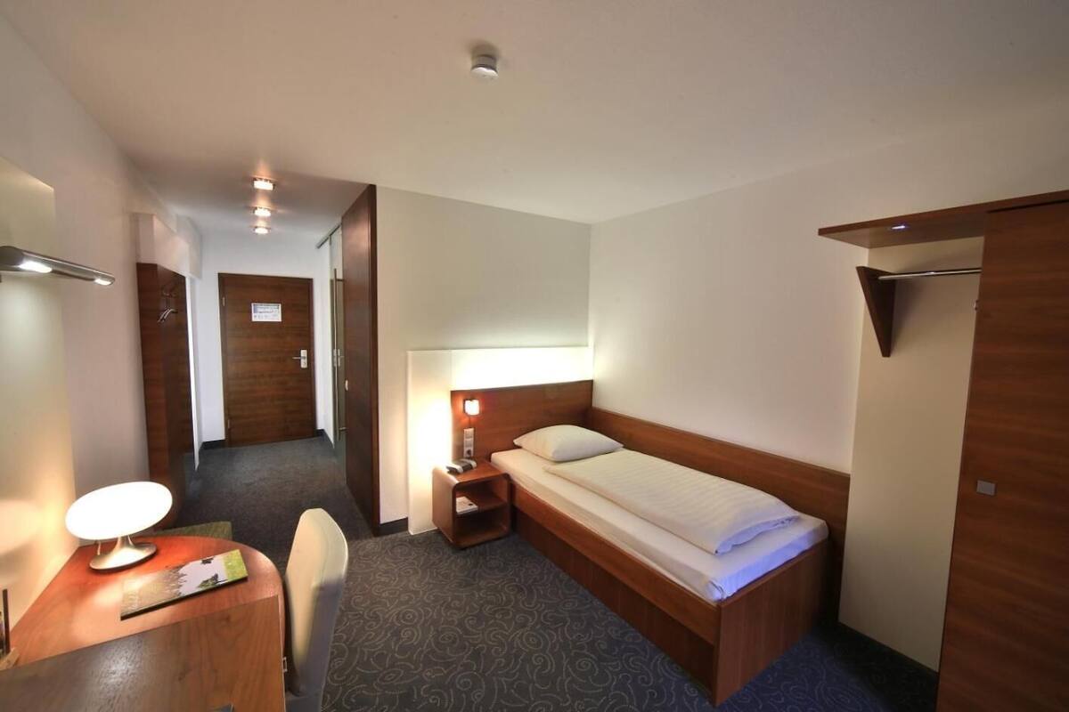 Ein Hotelzimmer mit Bett, Nachttisch, Lampe, Sessel und Schreibtisch mit Lampe.
