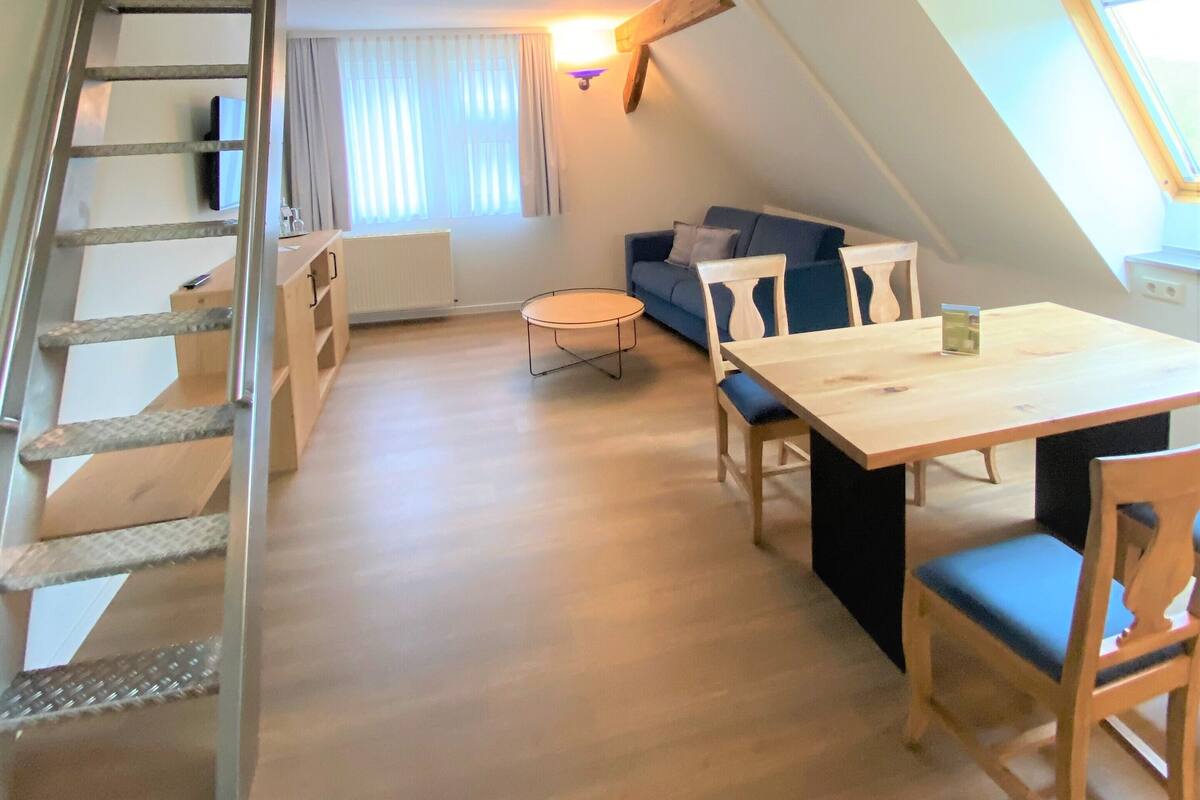 Eine Loft-artige Wohnung mit einer Holztreppe, einem Essbereich mit Tisch und Stühlen, einem Sofa und einem Fenster mit Vorhängen.