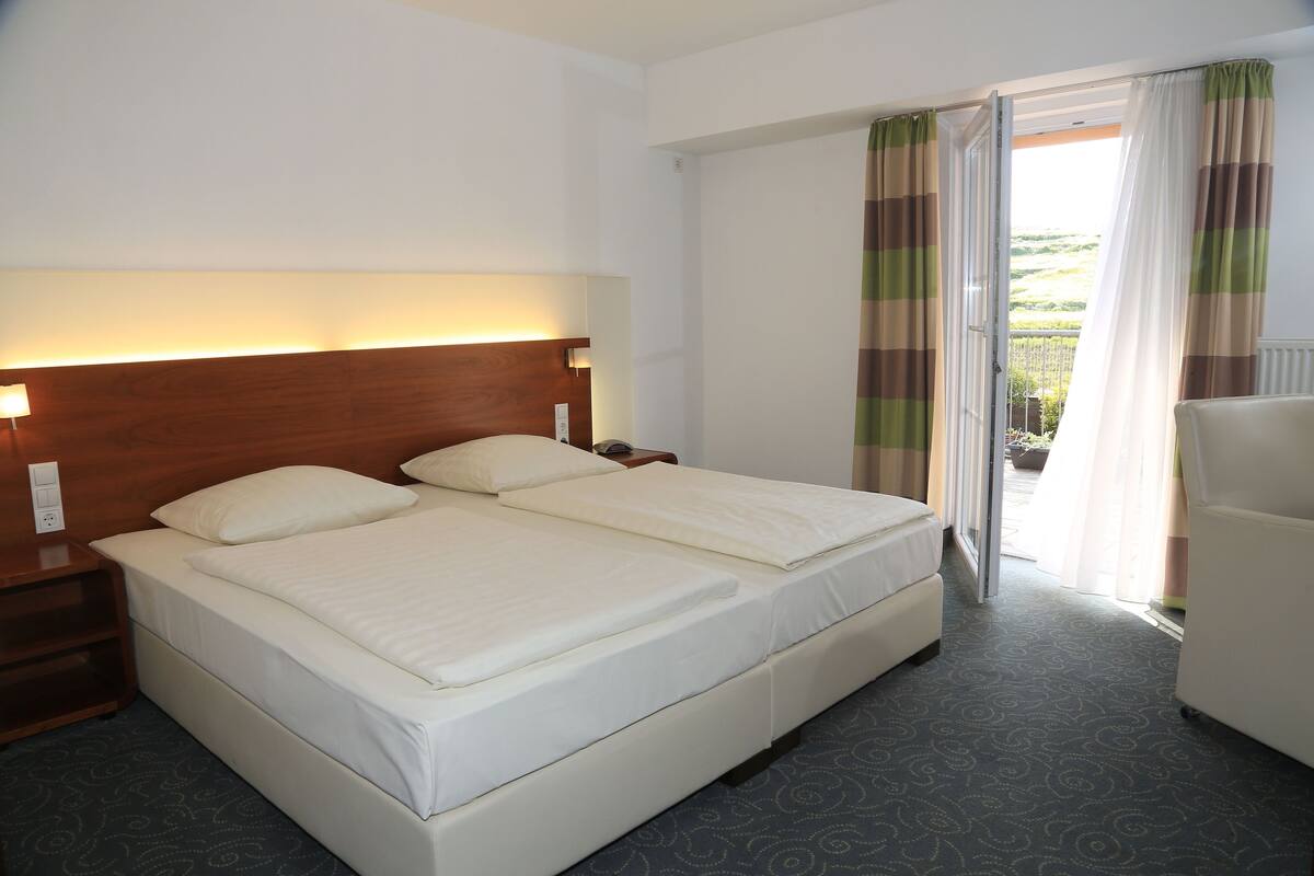 Ein Hotelzimmer mit einem großen Bett, einem Nachttisch, einer Lampe und Blick ins Freie durch ein Fenster mit Vorhängen.