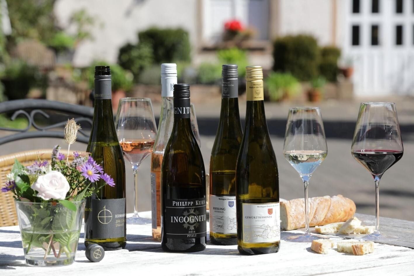 Una mesa con botellas de vino y copas, una cesta de flores y una hogaza de pan.