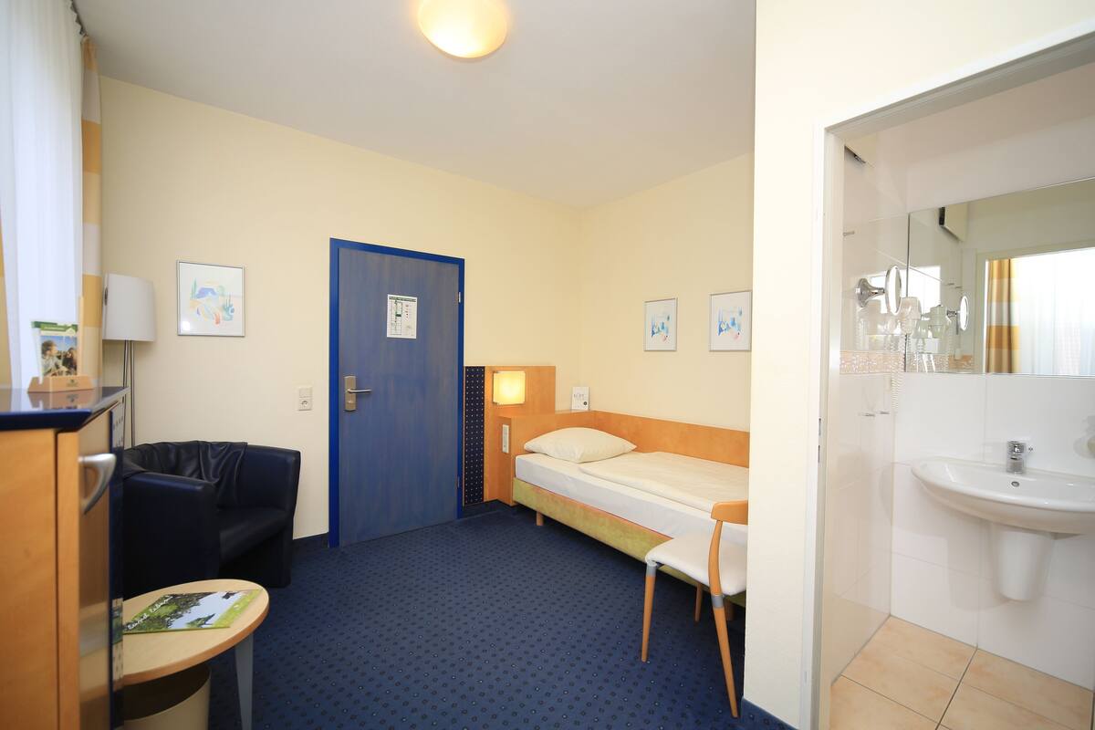 Ein Hotelzimmer mit einem Einzelbett, einem Schreibtisch mit Stuhl, einem kleinen Tisch, einer Sitzecke und einem Badezimmer mit Waschbecken und Spiegel.