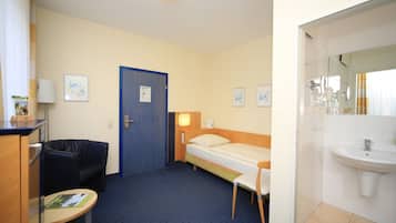 A hotel room with a single bed, a desk with a chair, a small table, a sofa, and a bathroom with a sink and mirror.