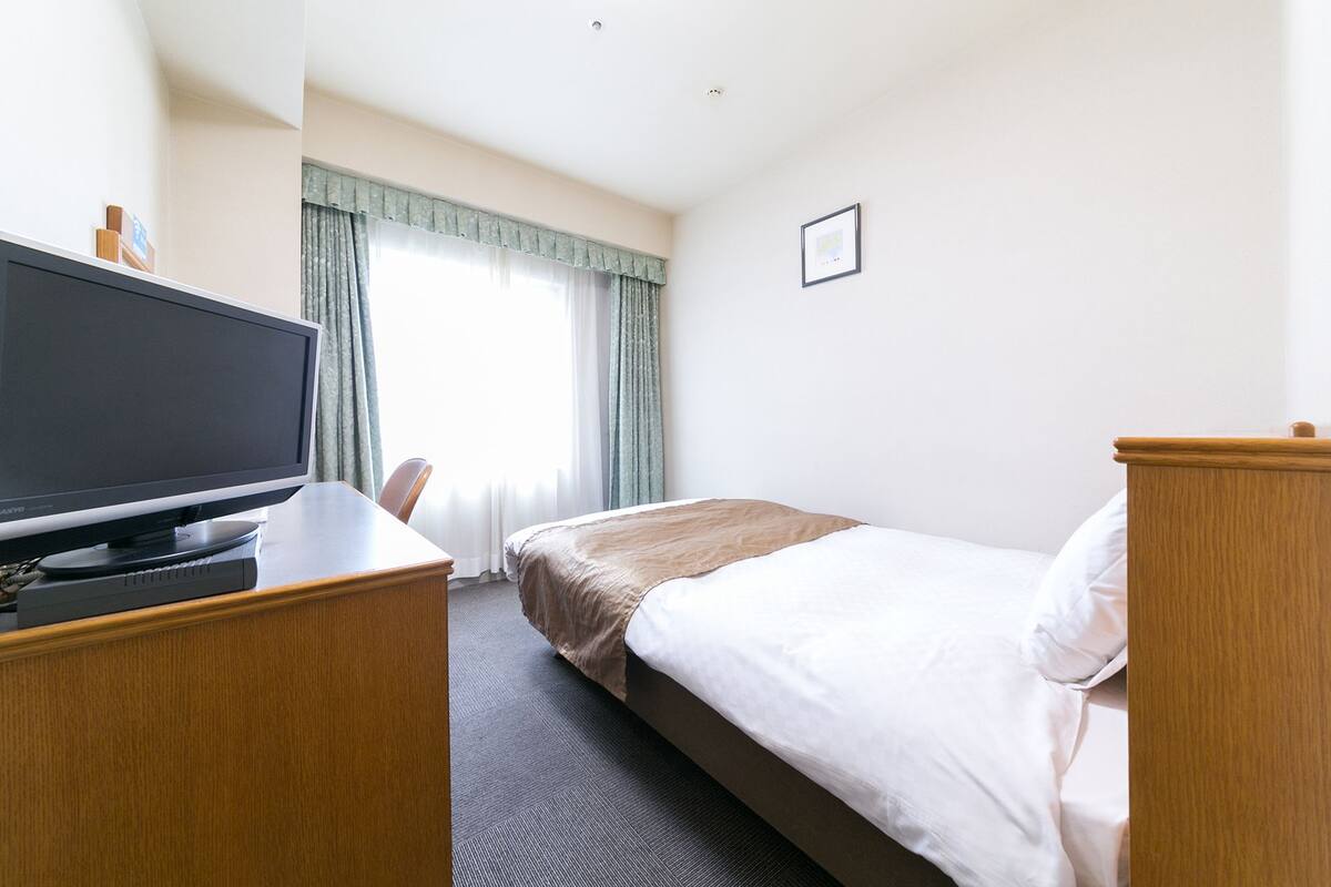 A hotel room with a bed, a desk with a television, and a window with curtains.