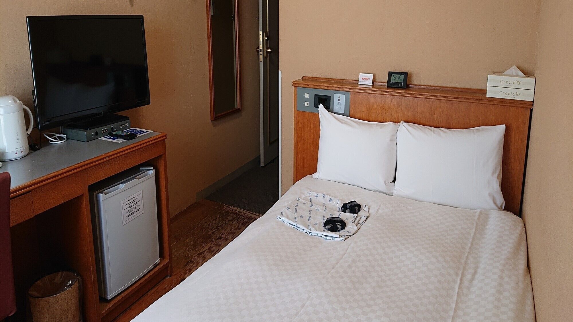 A hotel room with a bed, a desk with a television, a small refrigerator, and a bedside table with a clock and a tissue box.