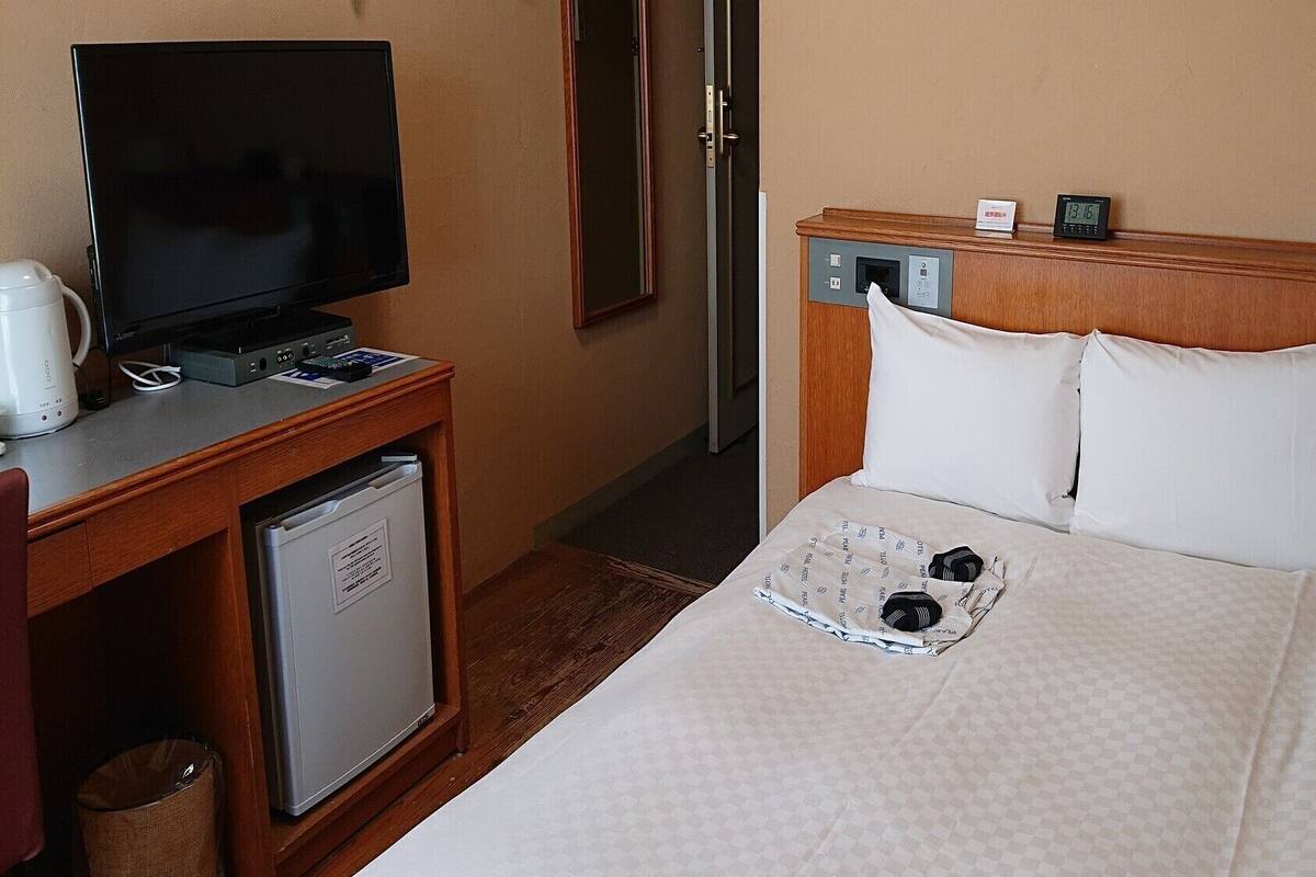 A hotel room with a bed, a desk with a television, a small refrigerator, and a bedside table with a clock and a tissue box.