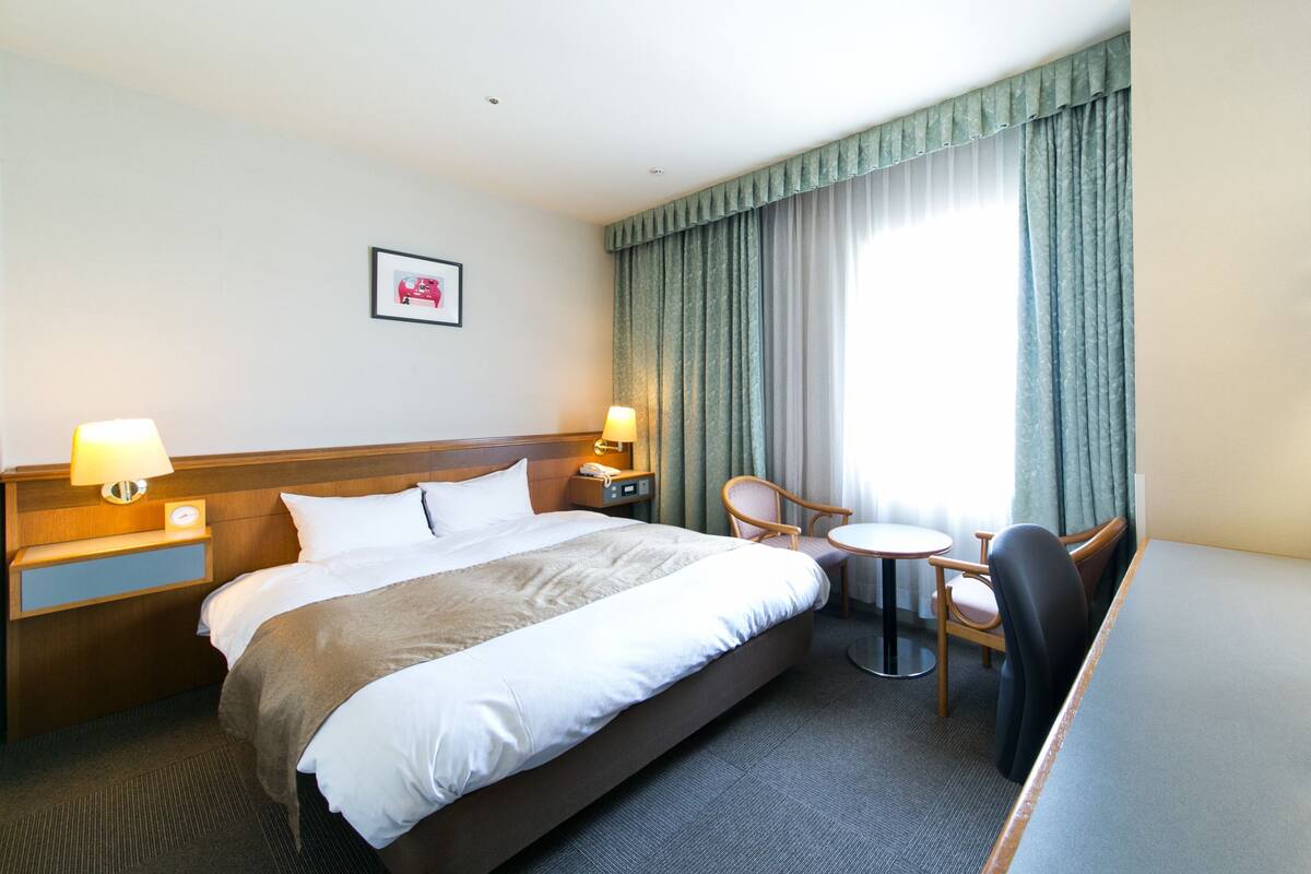 A hotel room with a large bed, bedside tables with lamps, a small round table, a chair, and a window with curtains.