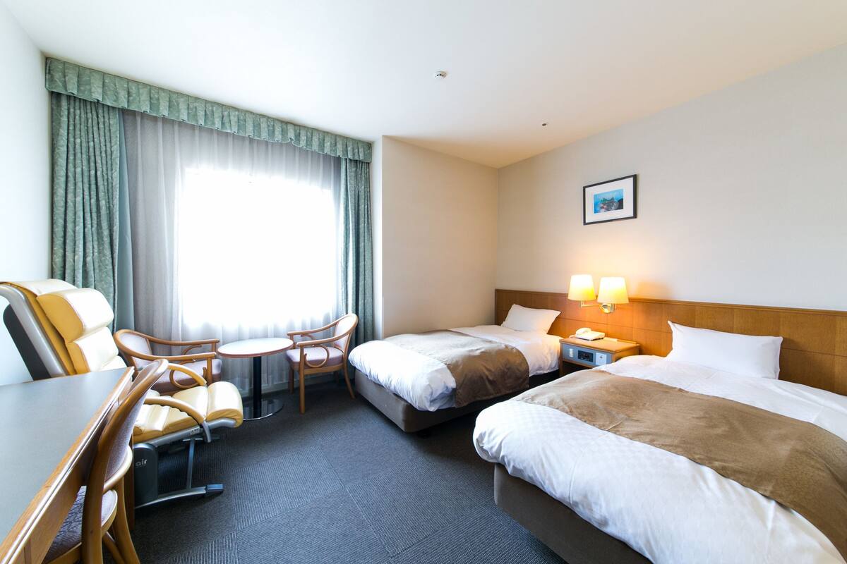 A hotel room with two beds, a desk with a chair, a small table, and a window with curtains.