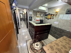 A hotel reception area with a reception desk, a wall-mounted calendar, an air conditioning unit, and a patterned rug.