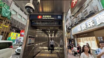 An indoor escalator leading to a station with signage in a foreign language, surrounded by commercial buildings and parked cars.