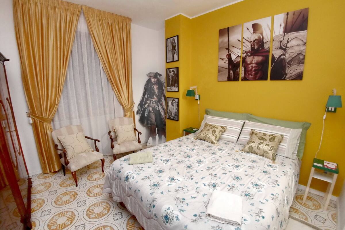 A bedroom with a bed, two armchairs, a nightstand, and a wall with framed pictures.