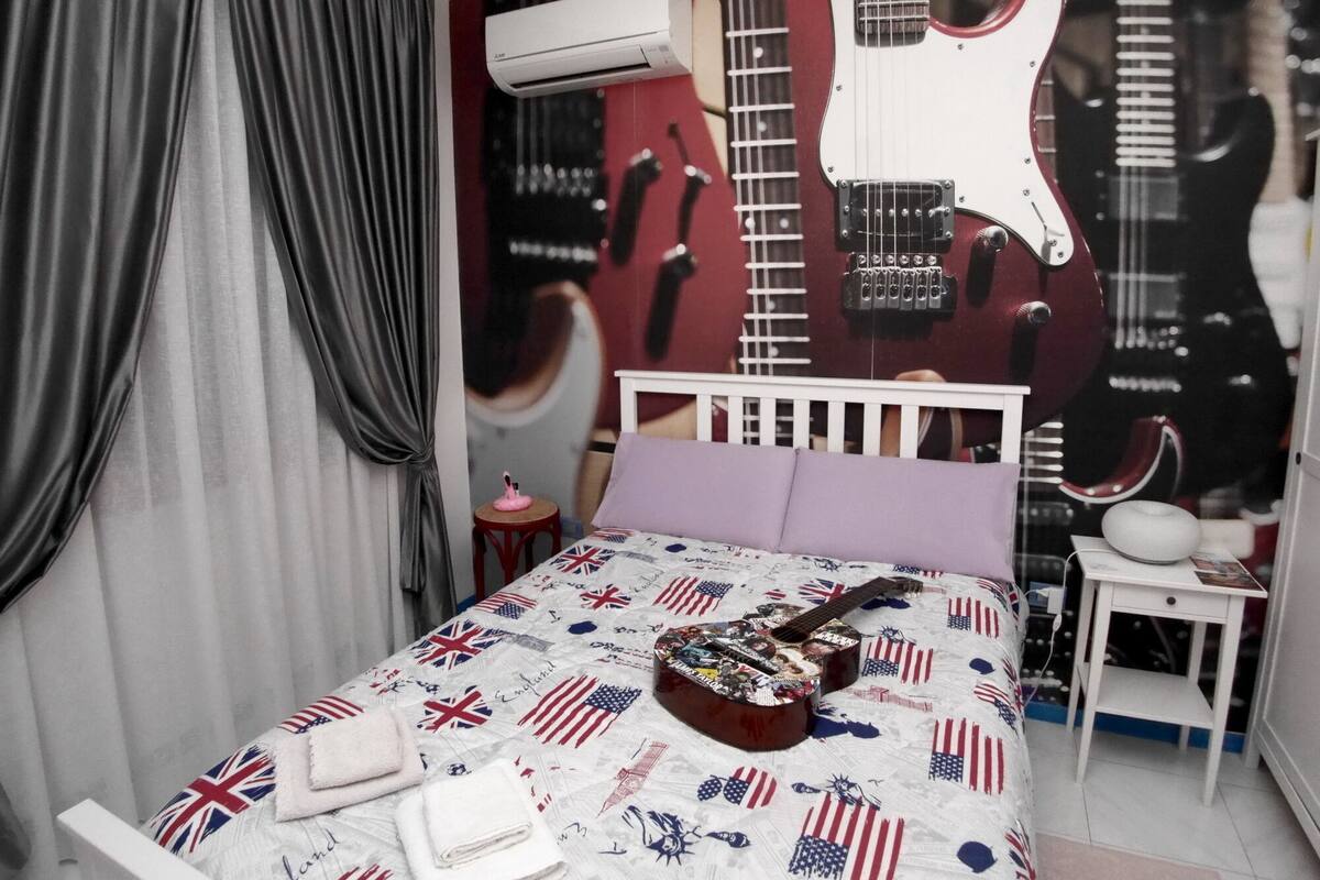 A bedroom with a bed, a guitar, a small table, and an air conditioning unit.