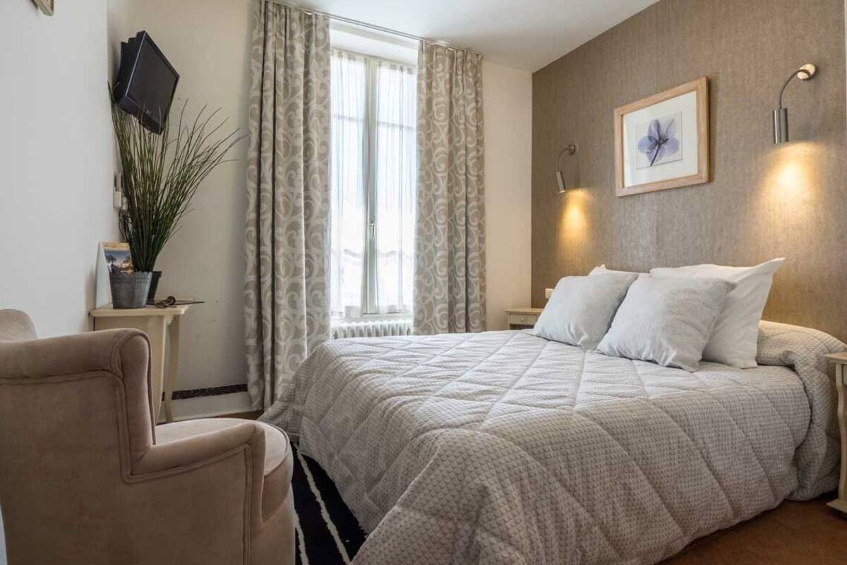 Un dormitorio con cama, una silla, un ventanal con cortinas, un cuadro en la pared y una lámpara de noche.