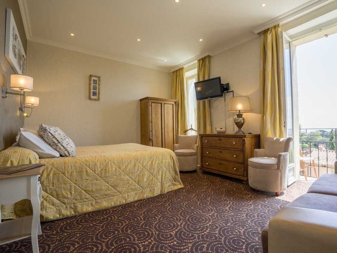 A hotel room with a bed, a desk, a dresser, a TV, and a balcony with yellow curtains.