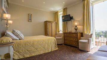 A hotel room with a bed, a desk, a dresser, a TV, and a balcony with yellow curtains.