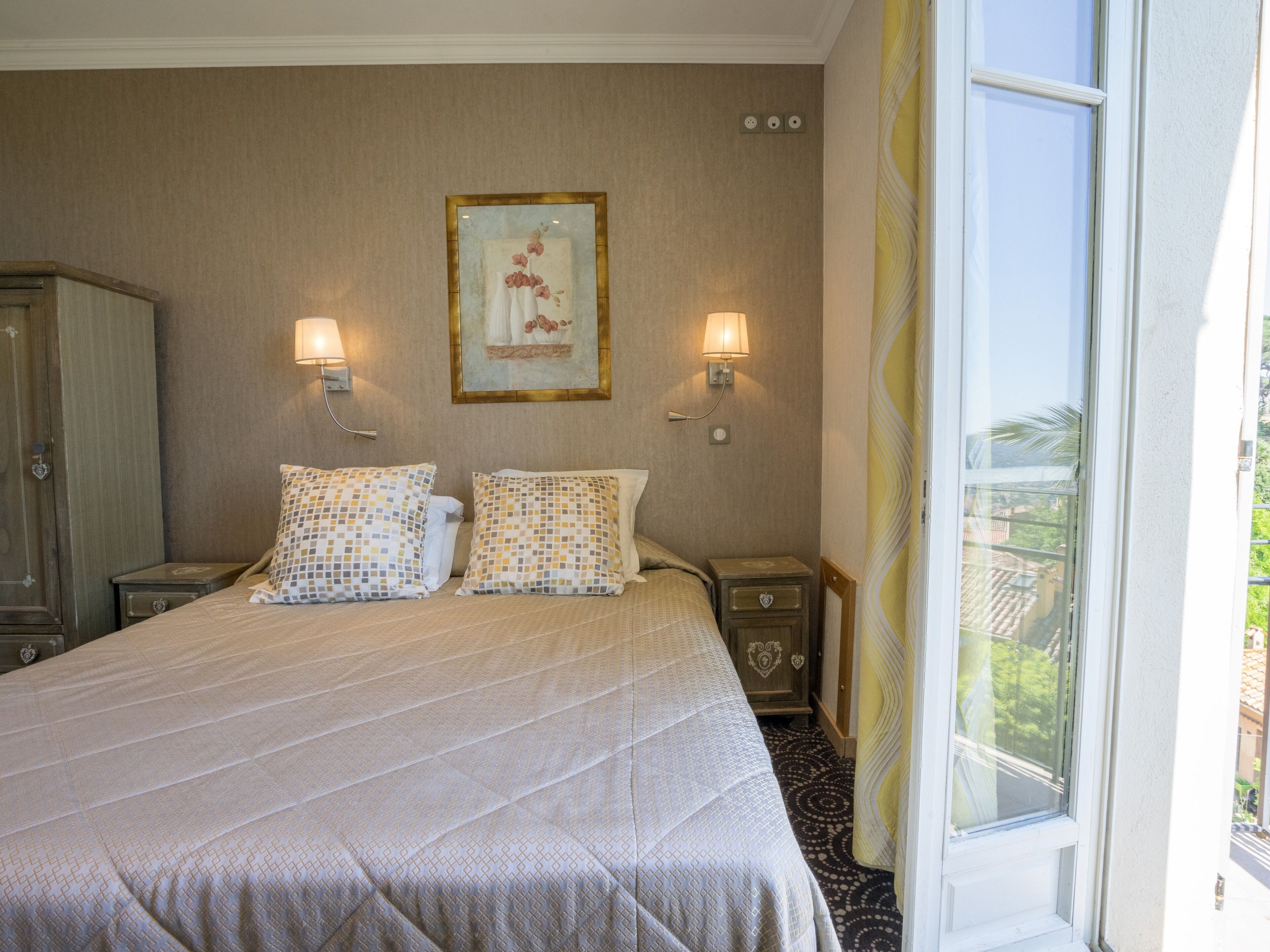 A hotel room with a large bed, two bedside tables with lamps, a framed artwork on the wall, and a window with a view of greenery.