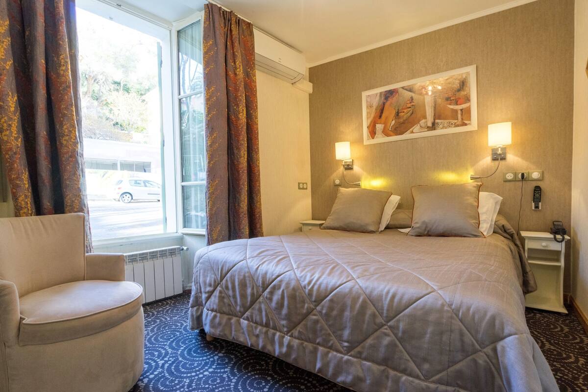 Una habitación de hotel con una cama, dos mesitas de noche, una silla, un ventanal con cortinas y un cuadro en la pared.