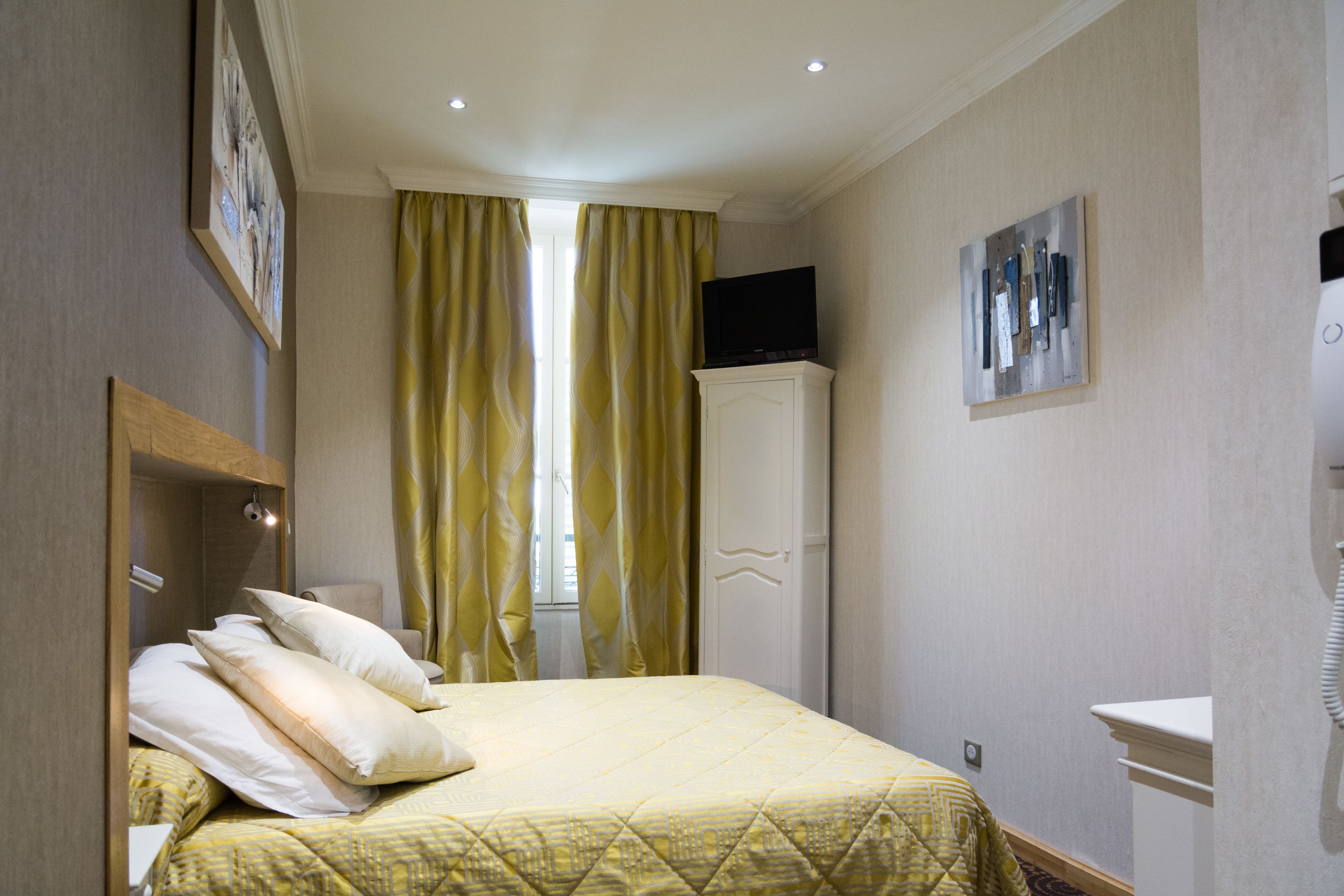 Une chambre d’hôtel avec un lit, un téléviseur posé sur une commode, un tableau accroché au mur et une fenêtre avec des rideaux.