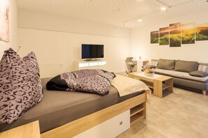 Una sala moderna con un sofá, una cama con almohadones estampados, un televisor y una pared con un cuadro de paisaje.