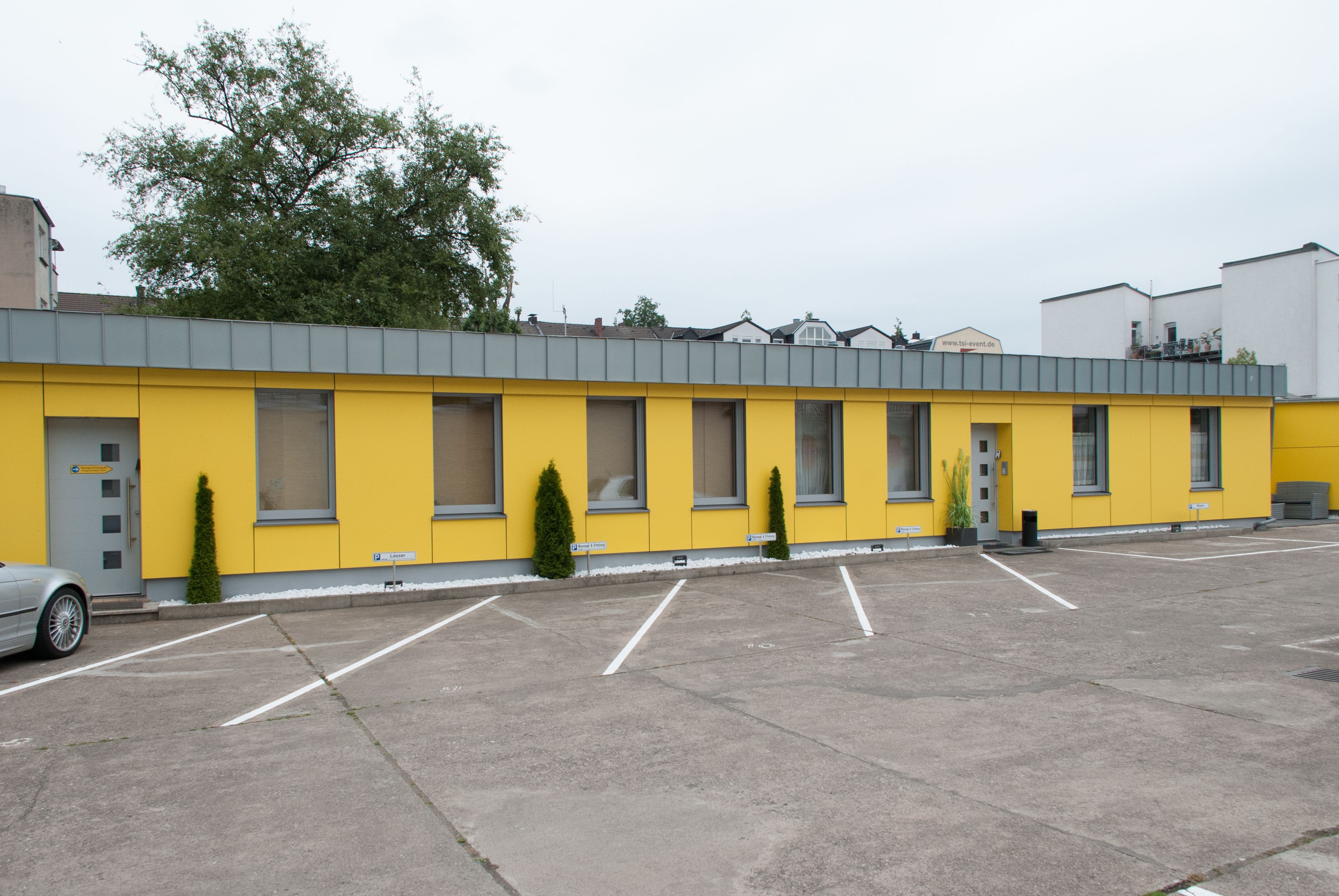 A yellow building with a grey roof, a parking lot, and a tree in the background.
