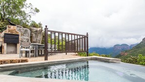 Une piscine bordée d’un mur en pierre et entourée d’une clôture en bois, le tout noyé dans un écrin de verdure et dominé par un paysage montagneux.