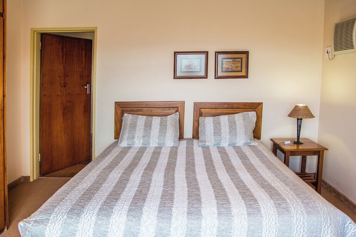 A double bed with striped bedding, two framed pictures on the wall, a bedside lamp, and a wooden door.