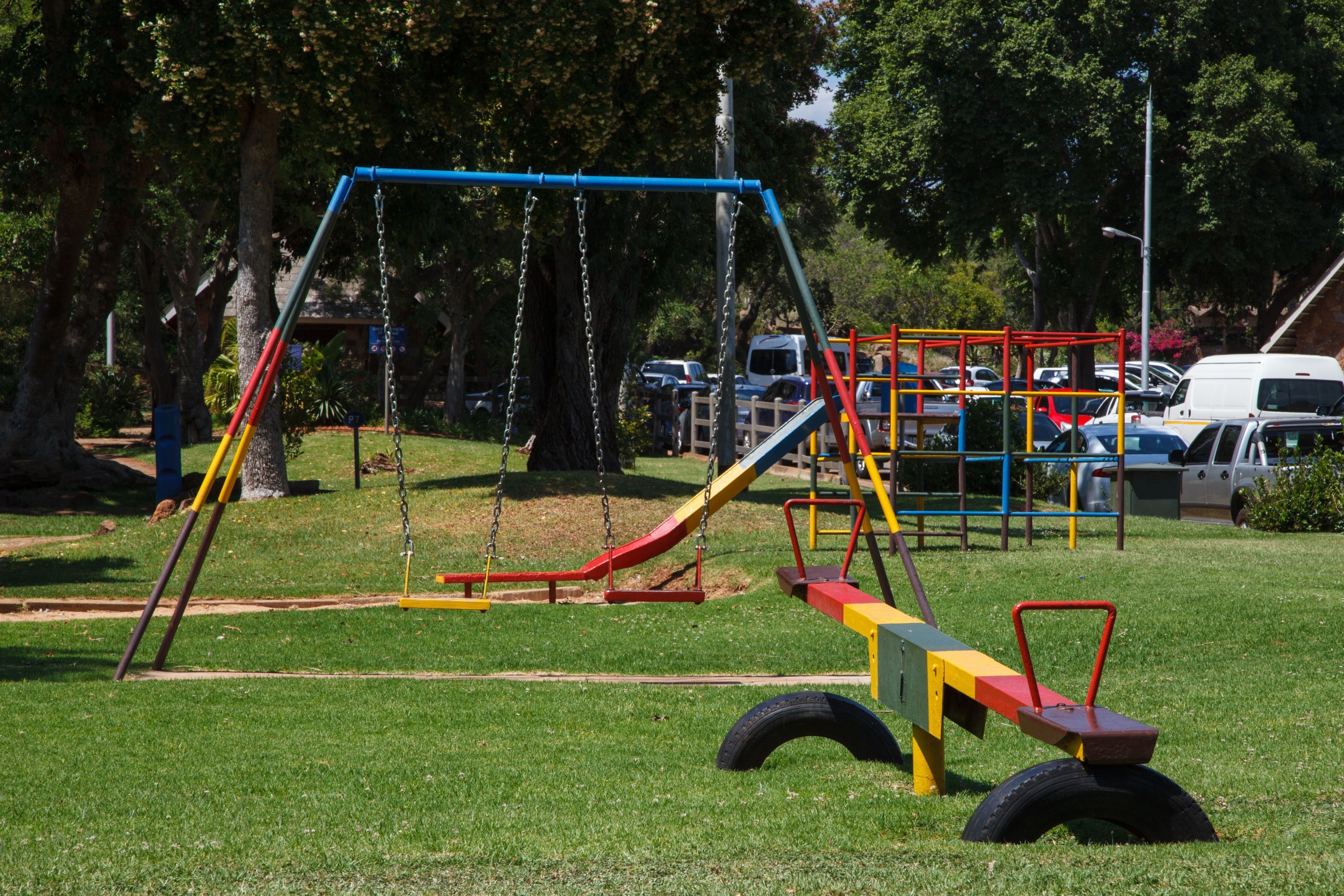 A playground with swings and slides, surrounded by trees and parked cars.