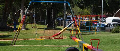 Un parque infantil con columpios y toboganes, rodeado de árboles y coches aparcados.