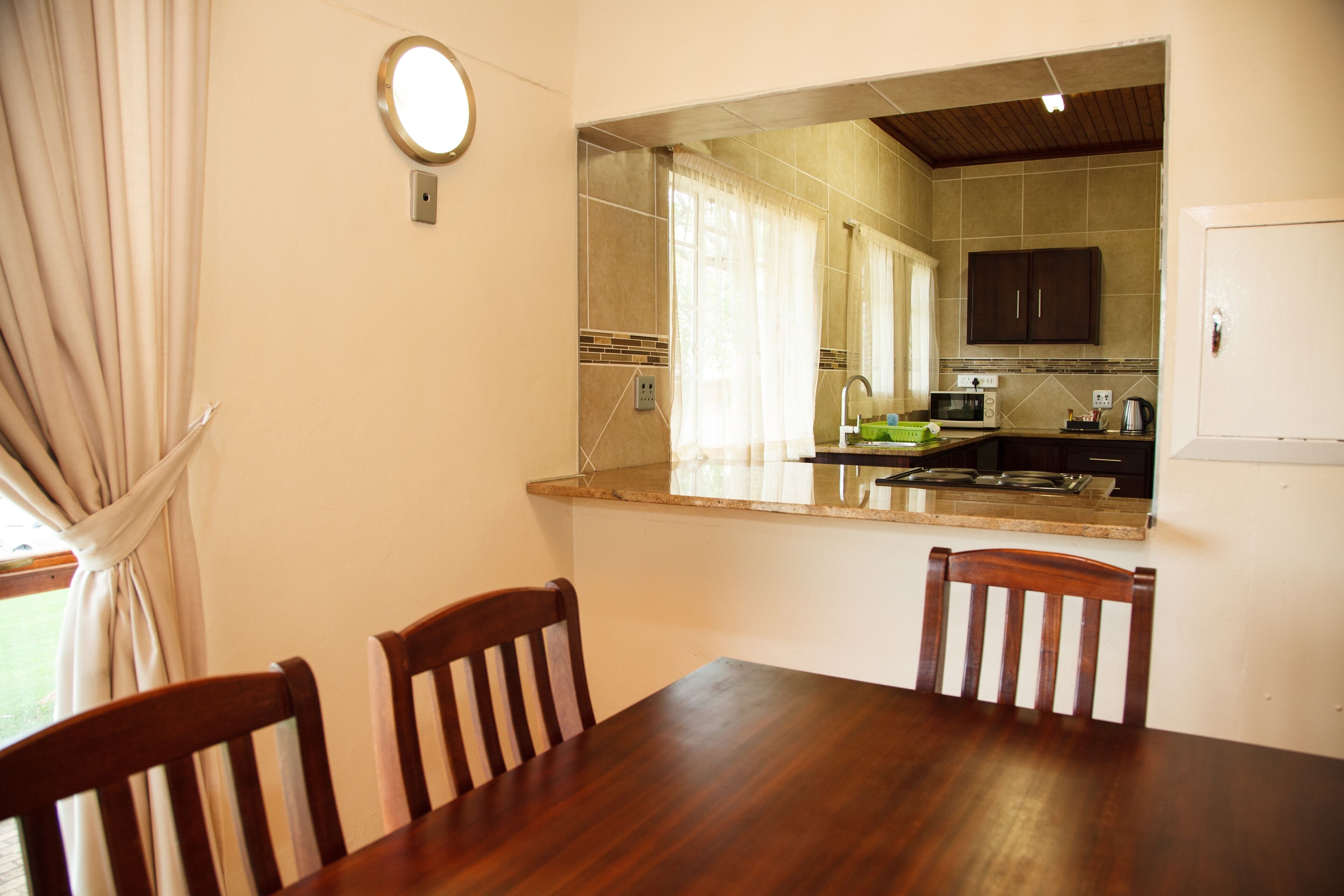 A dining area with a wooden table and chairs, a kitchen visible through a window, and a white door.